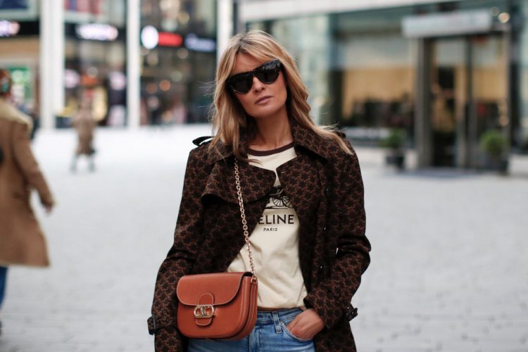 DUESSELDORF, GERMANY - DECEMBER 10: Influencer Gitta Banko wearing a dark brown trenchcoat with 70's triomphe print by Celine, a cream colored logo print t-shirt by Celine, a cognac colored chain bag with gold  triomphe details by Celine, light blue slim fit denim jeans by AG Jeans and sunglasses by Bottega Veneta during a street style shooting on December 10, 2020 at Breuninger in Duesseldorf, Germany. (Photo by Streetstyleshooters/Getty Images)
